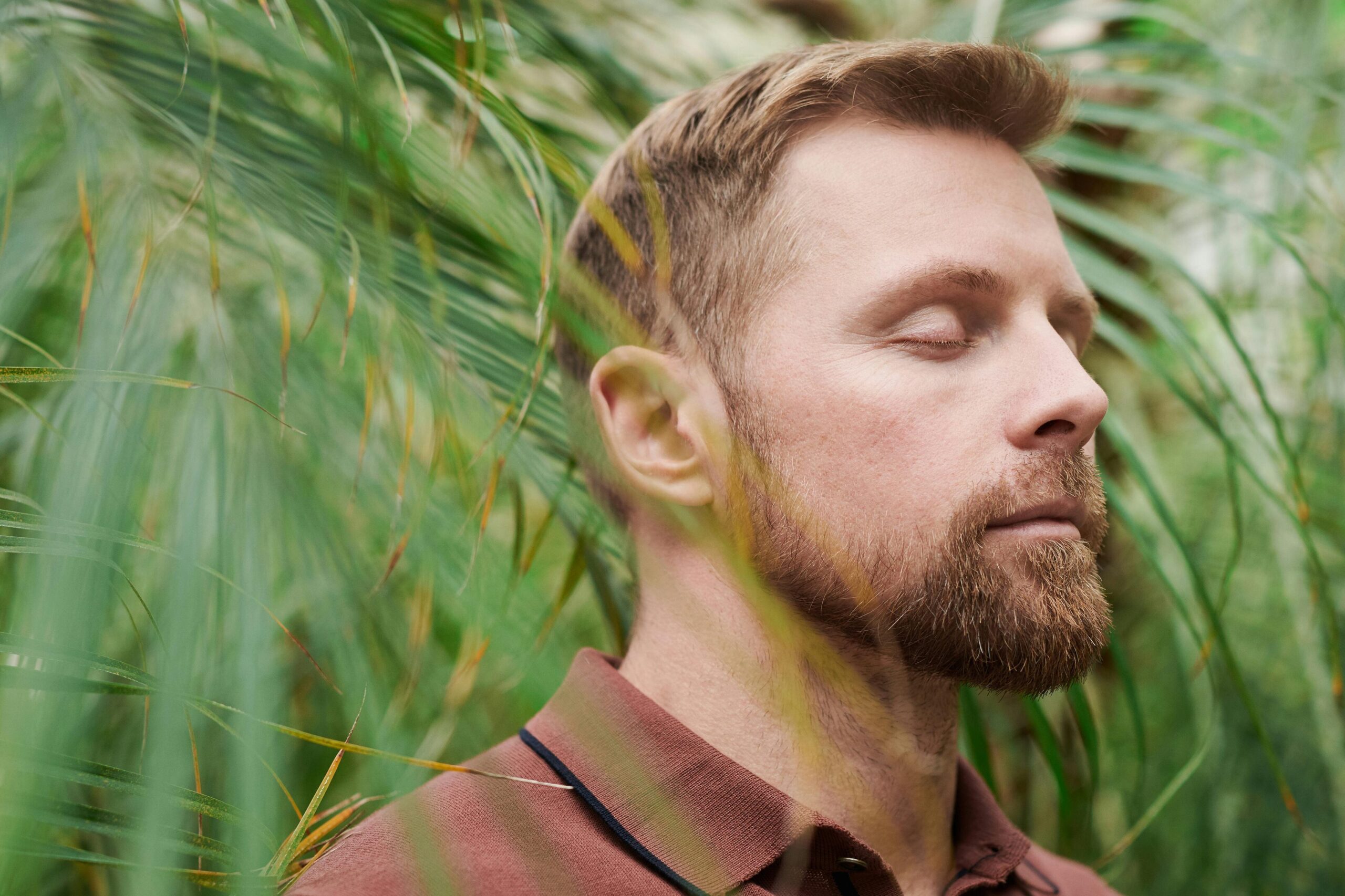 A man with closed eyes meditates peacefully amidst lush green palm leaves, promoting relaxation and mindfulness.