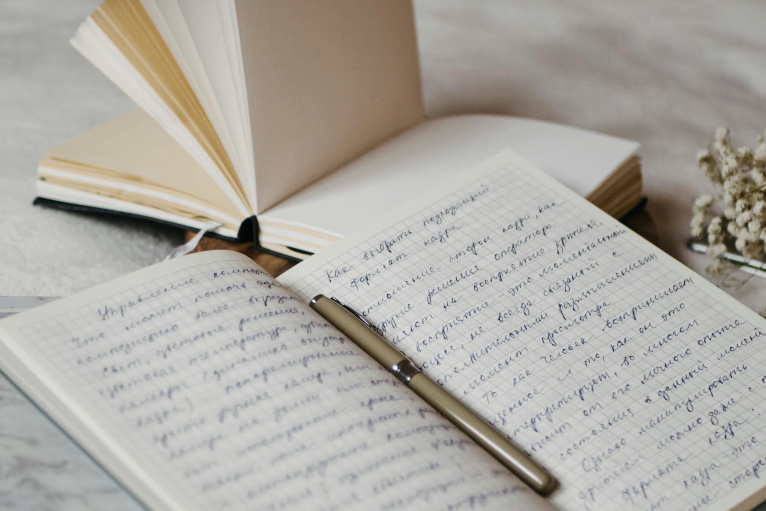 Close-up of open notebooks and a pen on a soft bed surface with delicate dried flowers.
