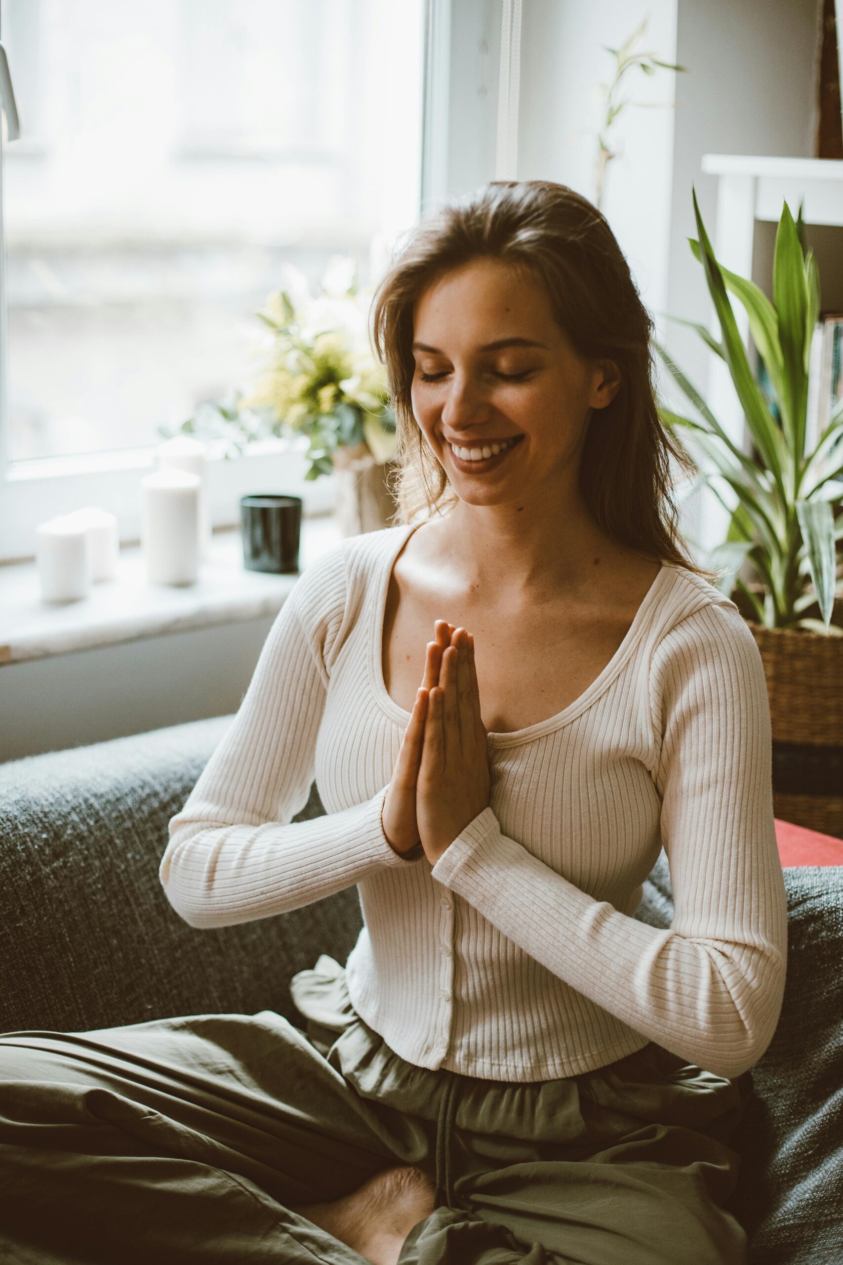 Portrait of a smiling woman meditating on a sofa with hands in prayer position.