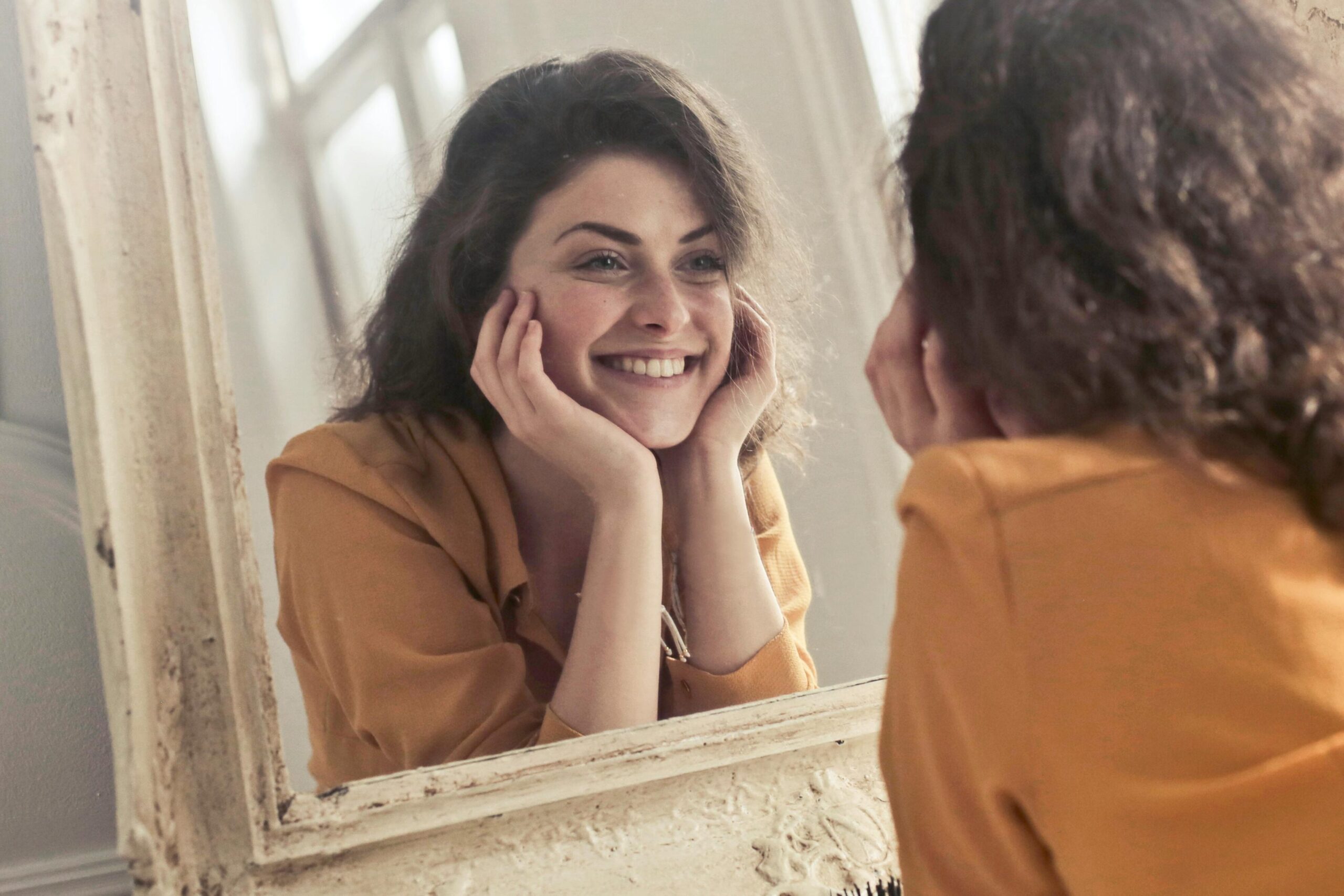A cheerful woman smiles at her reflection in a vintage-style mirror, exuding positivity and warmth.